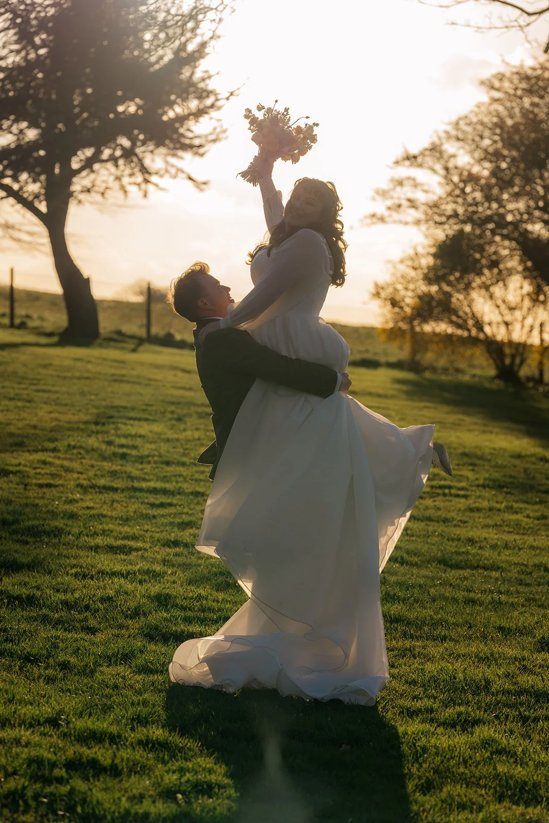 Josh lifts Emily into the air during a playful golden-hour wedding portrait at Coombe Lodge in Somerset, UK, as she raises her bouquet against the sunset.