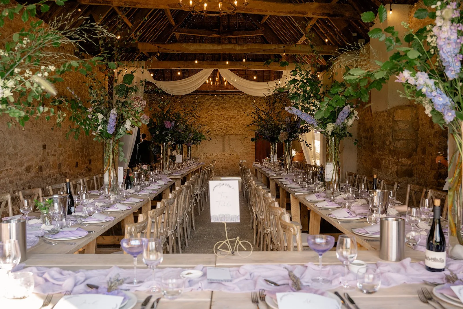 Indoor wedding breakfast setup at The Cider Barn in Somerset for Abi & Josh’s wedding, with long rustic tables, lilac details, flowers and stone barn walls, photographed by Just James Weddings for relaxed barn wedding decor.