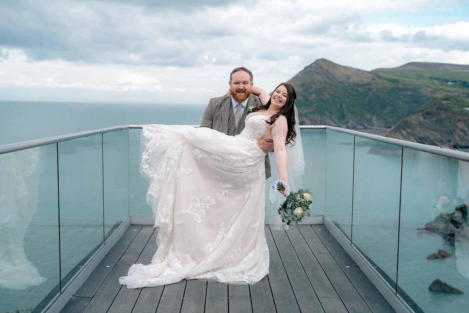 Terri &amp; Sam at Sandy Cove Hotel standing on a glass balcony with dramatic coastal cliffs behind them. Sam lifts Terri in his arms as her lace gown flows outward, both smiling against the backdrop of the sea and soft, cloudy sky.