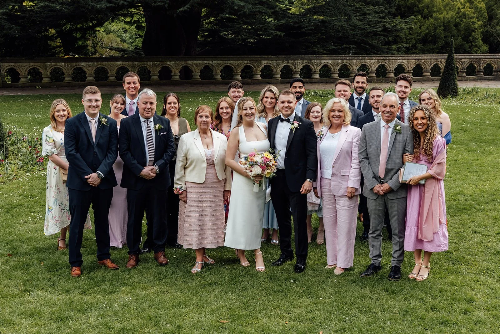 Bryony & Max stand with their wedding guests on the lawn at Insole Court in Cardiff, surrounded by greenery, pastel outfits and a decorative stone wall for a relaxed group portrait after their intimate ceremony and joyful confetti celebrations