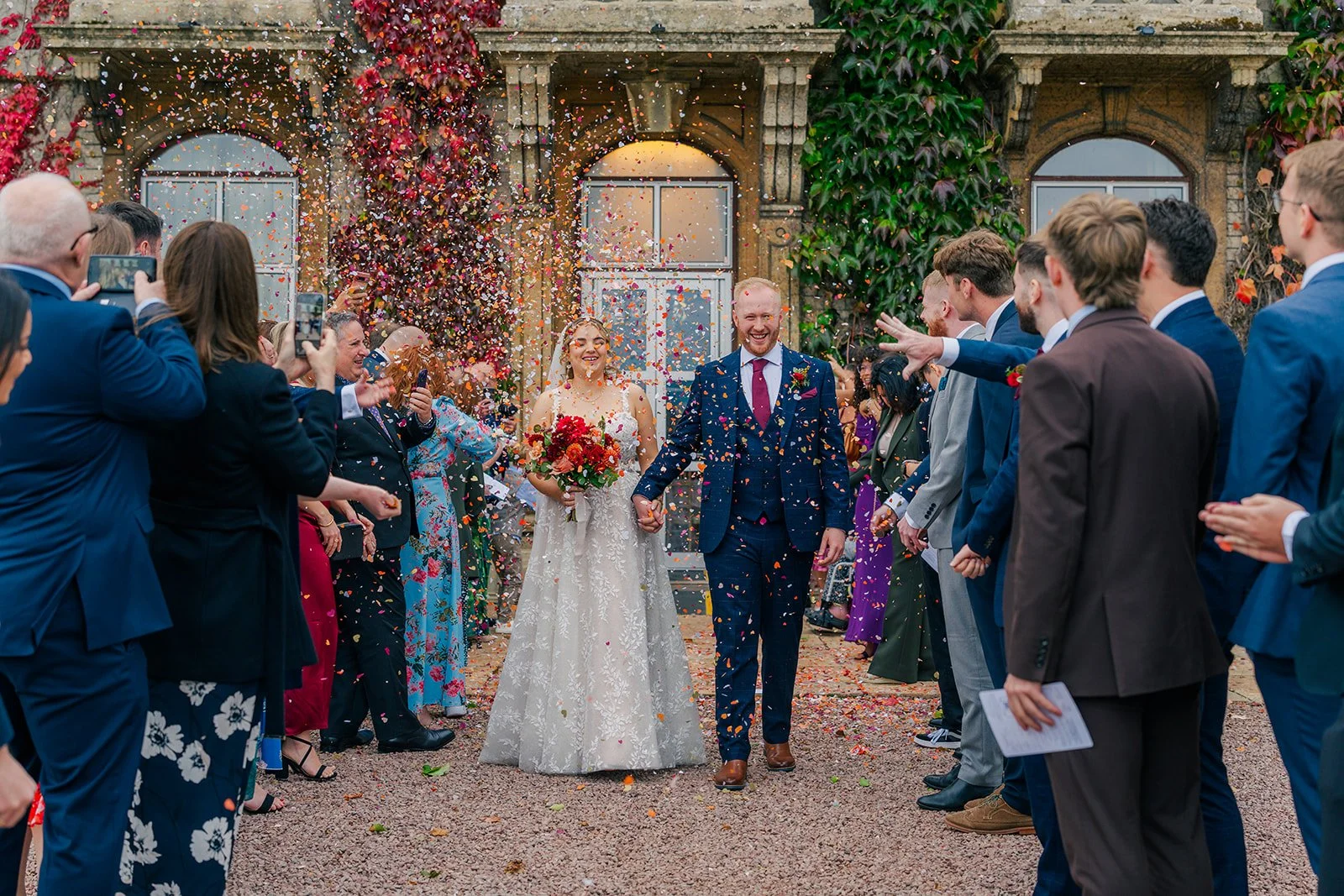 Lucy & Jay walking through colourful confetti at Eastwood Park in Gloucestershire, surrounded by cheering guests, photographed by Just James Weddings in a joyful documentary wedding style.
