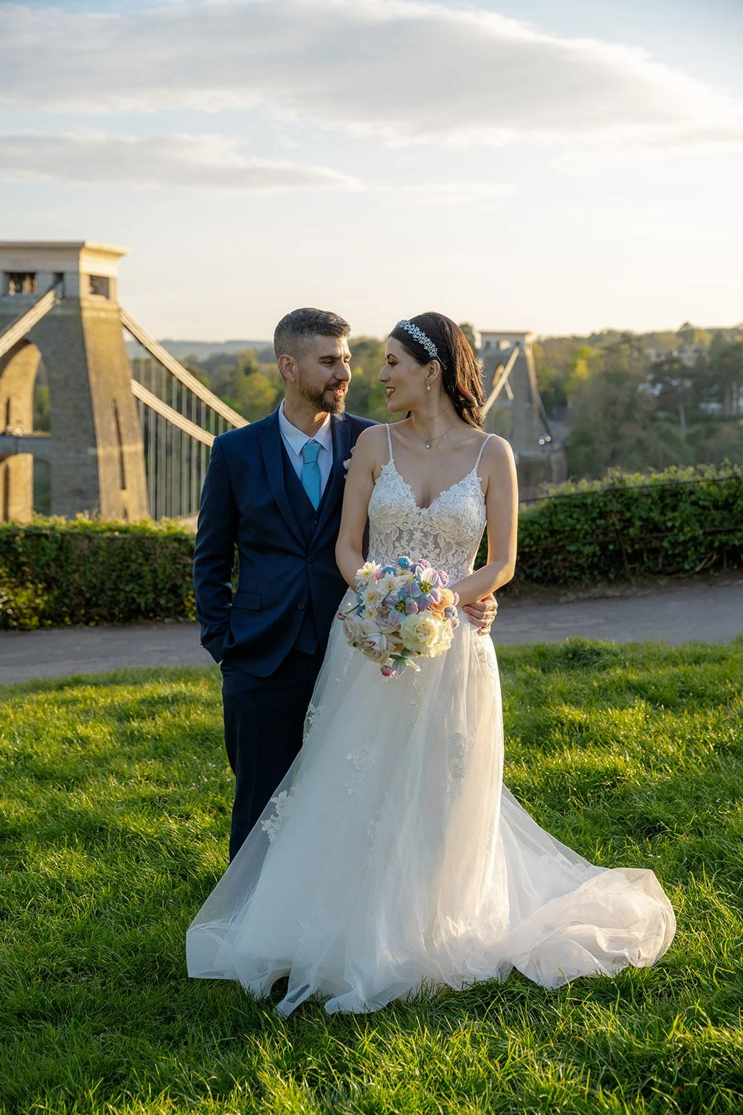 Ruth and Carmelo stand together on the grass with Clifton Suspension Bridge behind them in Bristol, UK, during their romantic wedding portraits after celebrating at No. 4 Clifton Village.