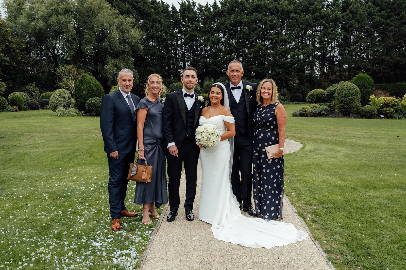 Nisha and Jack pose with family members on the landscaped gardens at Batch Country House in Somerset, UK, with Nisha holding her round white bouquet and Jack wearing a black tuxedo beside her, photographed by Just James Weddings.