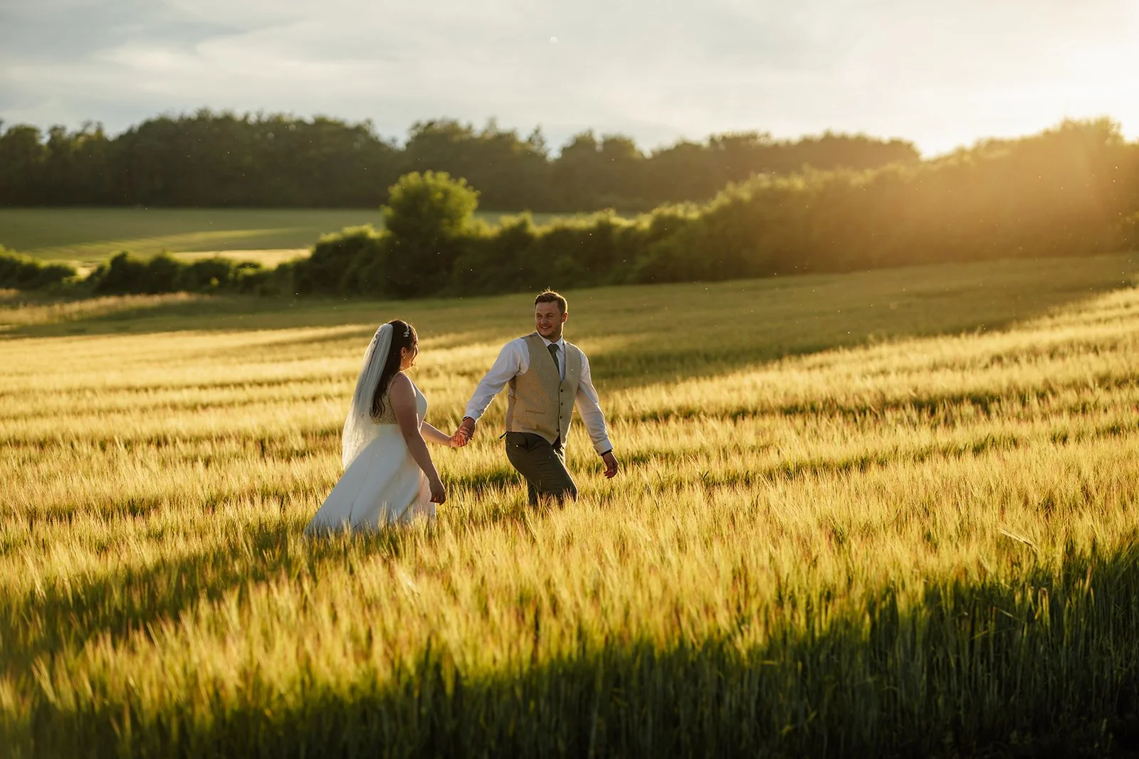 Chloe and Armani walk hand in hand through a sunlit field at golden hour near The Barn at Upcote in Gloucestershire, photographed by Just James Weddings.