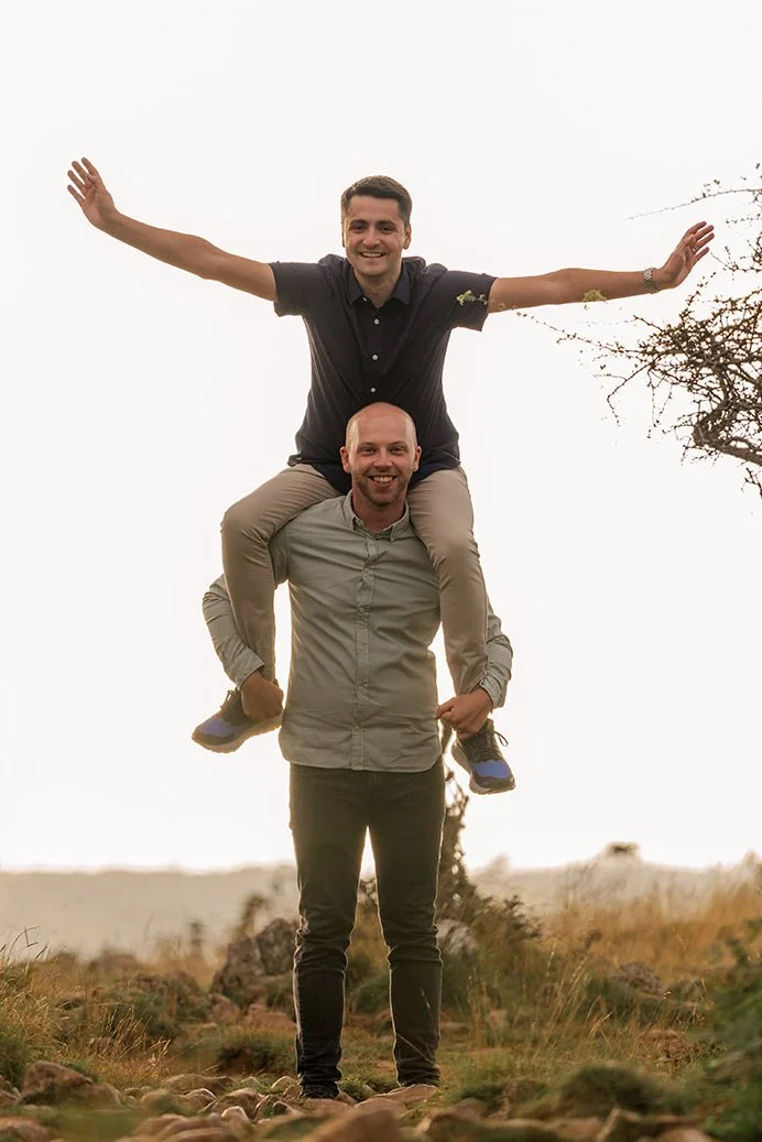 Sam sits on Gavin's shoulders with his arms spread wide and a huge grin during their engagement shoot on a rugged moorland hillside, with warm golden autumn light and wild grasses stretching out across the open landscape behind them.
