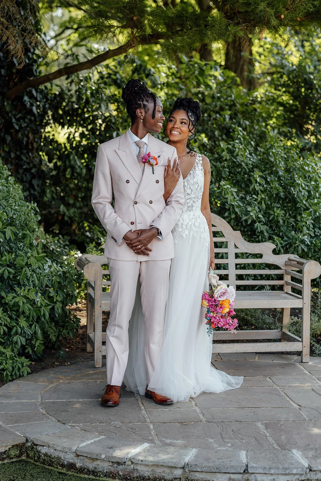 Kasey and Larisha smiling together beneath the trees during a relaxed couple portrait in the gardens at Oaks Farm Weddings in Surrey.