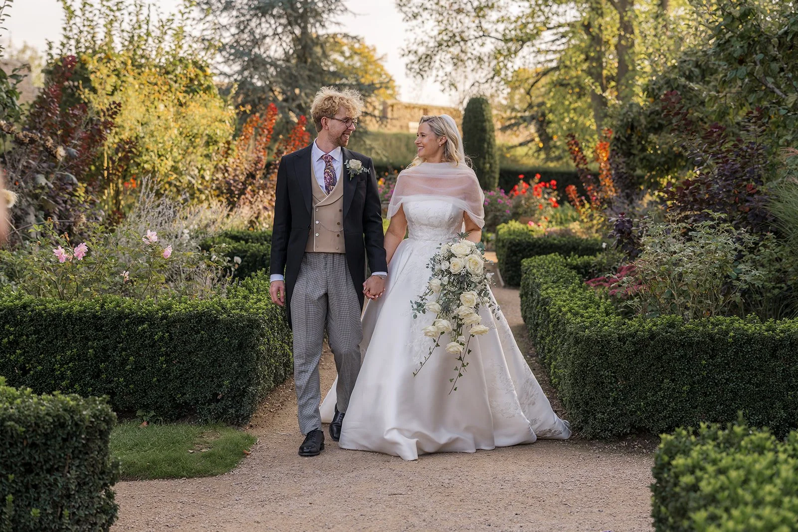 Iona & Will walking hand in hand through a manicured garden path on their wedding day at Iona & Will, somerset. The bride wears a classic white gown and carries cascading white roses as the groom looks at her smiling in soft golden light.