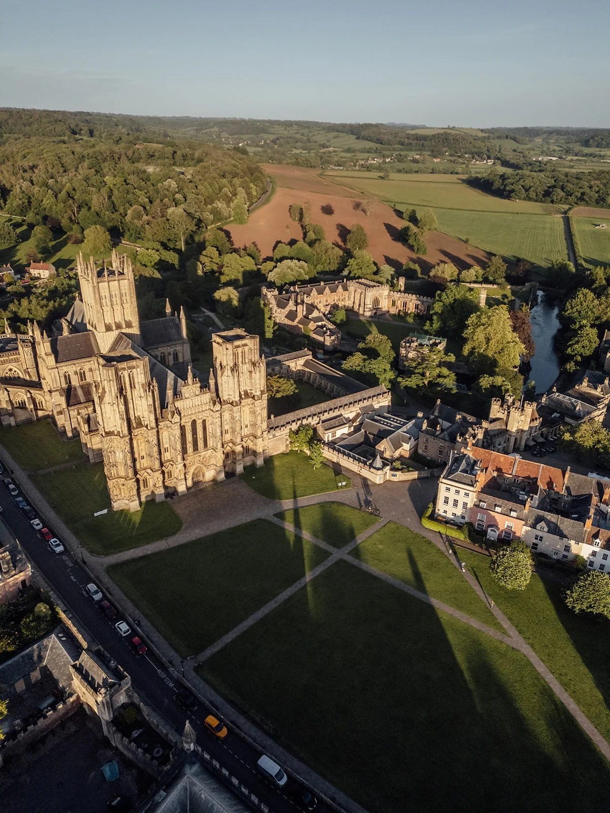 Aerial view of Charlotte & Jack’s wedding setting at Bishop’s Palace in Somerset, UK, showing Wells Cathedral, the palace buildings, gardens, river and surrounding countryside glowing in golden evening light after their wedding day in Wells.