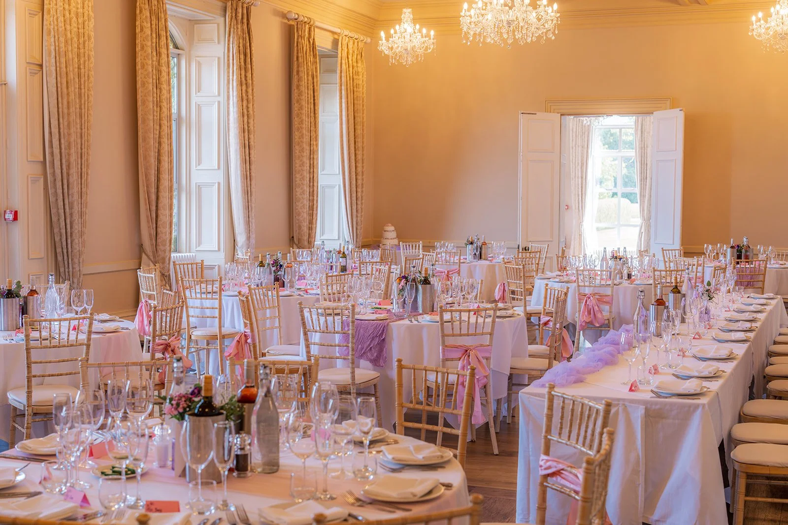 Wedding breakfast room at Eastwood Park in Gloucestershire, with long tables, gold chairs, pink details and chandeliers, photographed by Just James Weddings for reception decor inspiration.