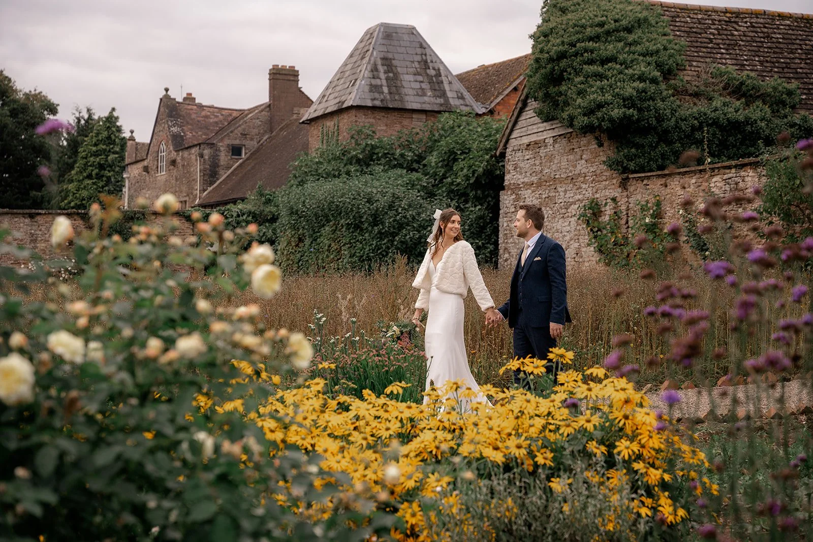Lilly & Lloyd walking hand in hand through the flower gardens at Broadfield Court in Herefordshire, surrounded by colourful blooms and historic buildings, photographed by Just James Weddings.