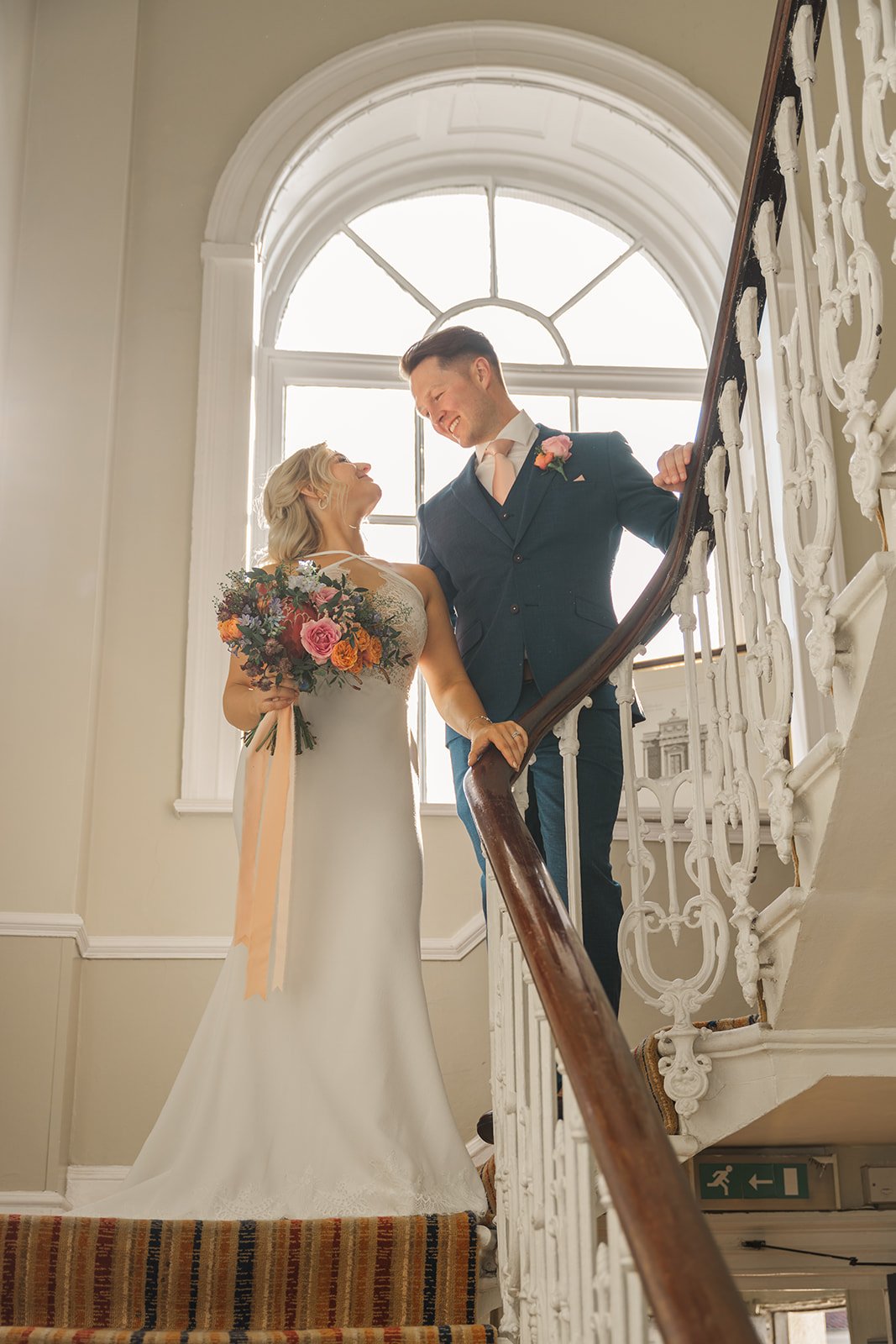 Kristy and Will smile at each other on the grand staircase at No. 4 Clifton Village in Bristol, UK, with Kristy holding a colourful wedding bouquet beside a large arched window.