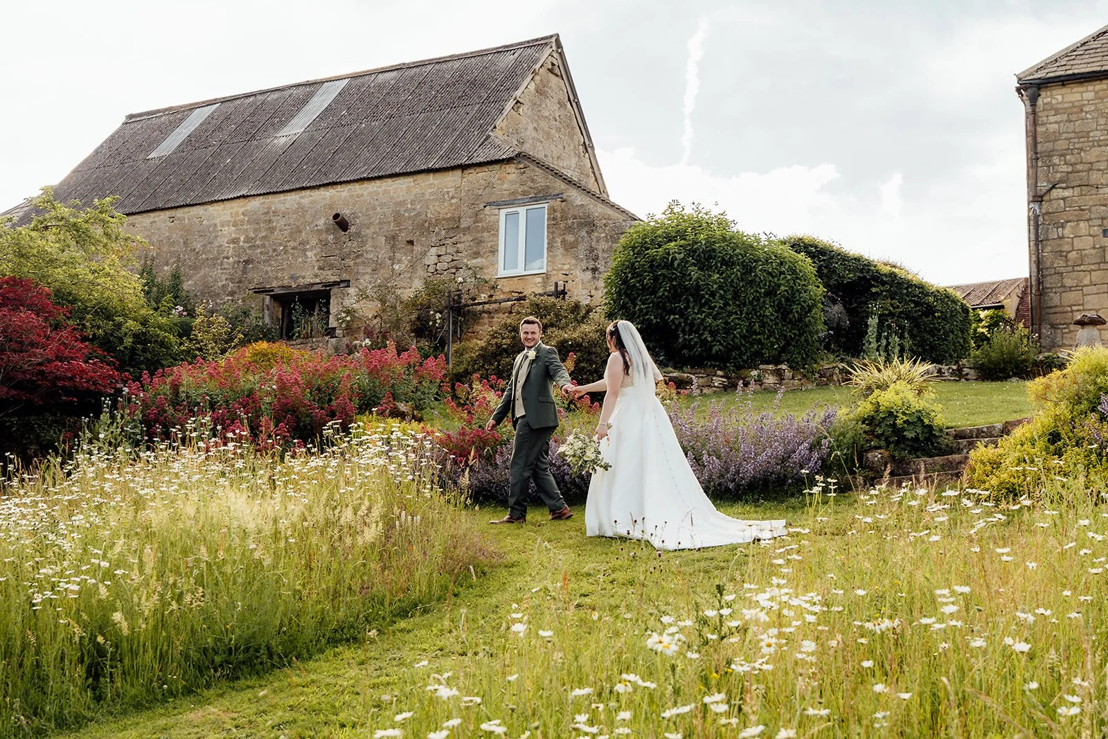 Chloe & Armani walking hand in hand through the gardens at The Barn at Upcote in the Cotswolds, surrounded by wildflowers and stone barns, photographed by Just James Weddings.