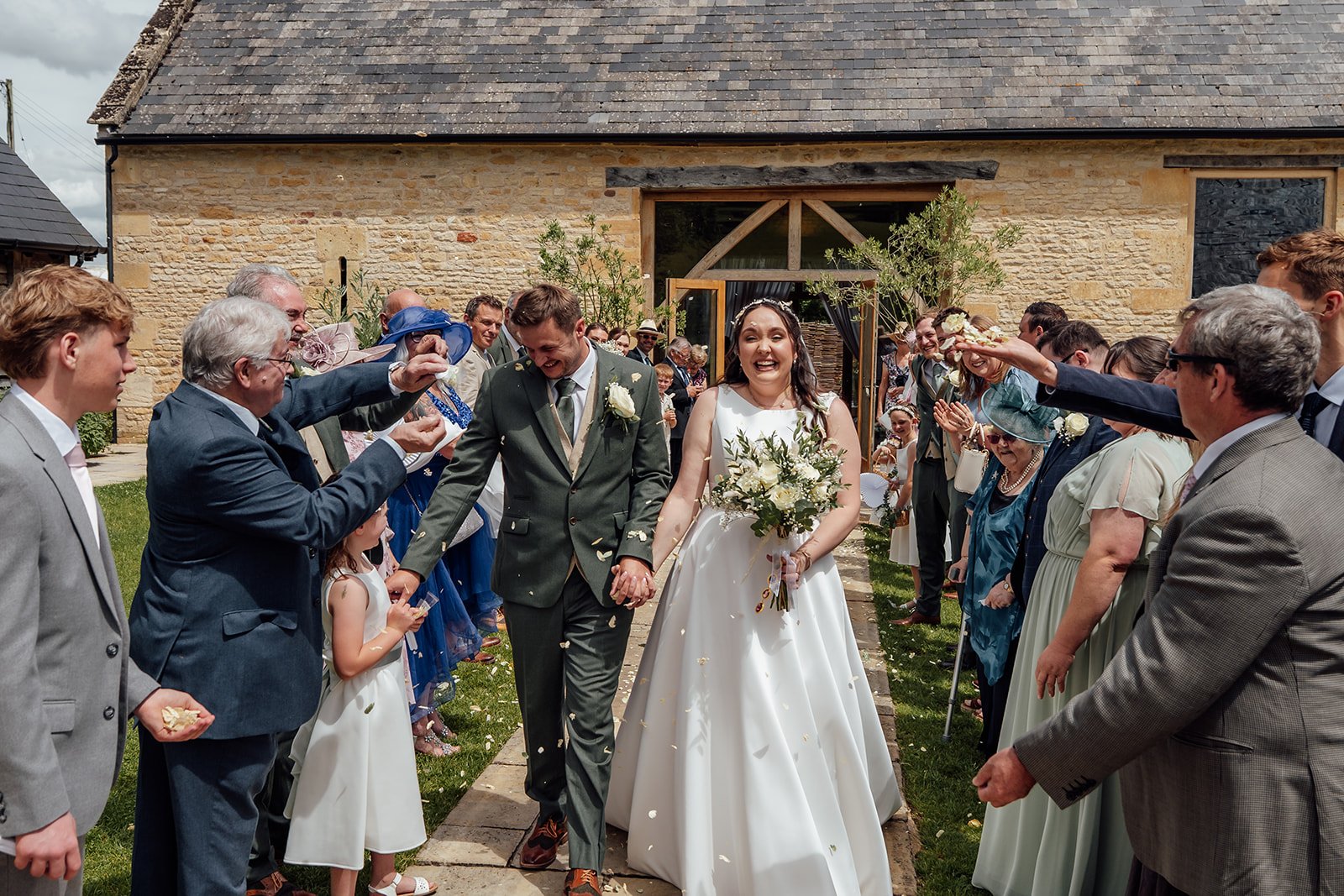 Chloe & Armani walking through confetti outside The Barn at Upcote in the Cotswolds, surrounded by cheering guests, photographed by Just James Weddings in a joyful documentary style.