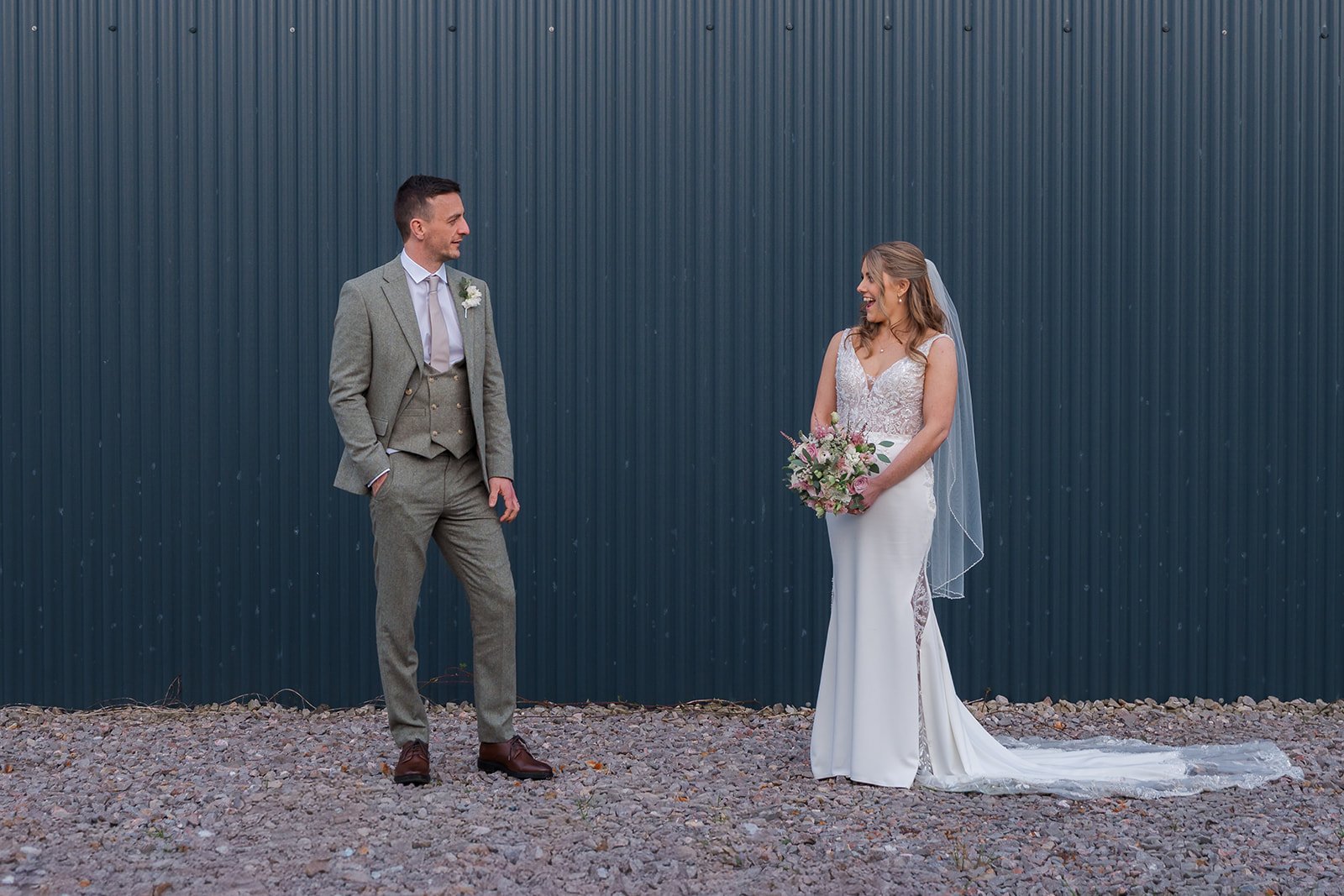 Anna and Tom stand apart laughing and sharing a candid moment together in front of the dark charcoal corrugated barn wall at Winkworth Farm in the Cotswolds, Anna holding her soft pink bouquet in her fitted lace wedding gown and veil.