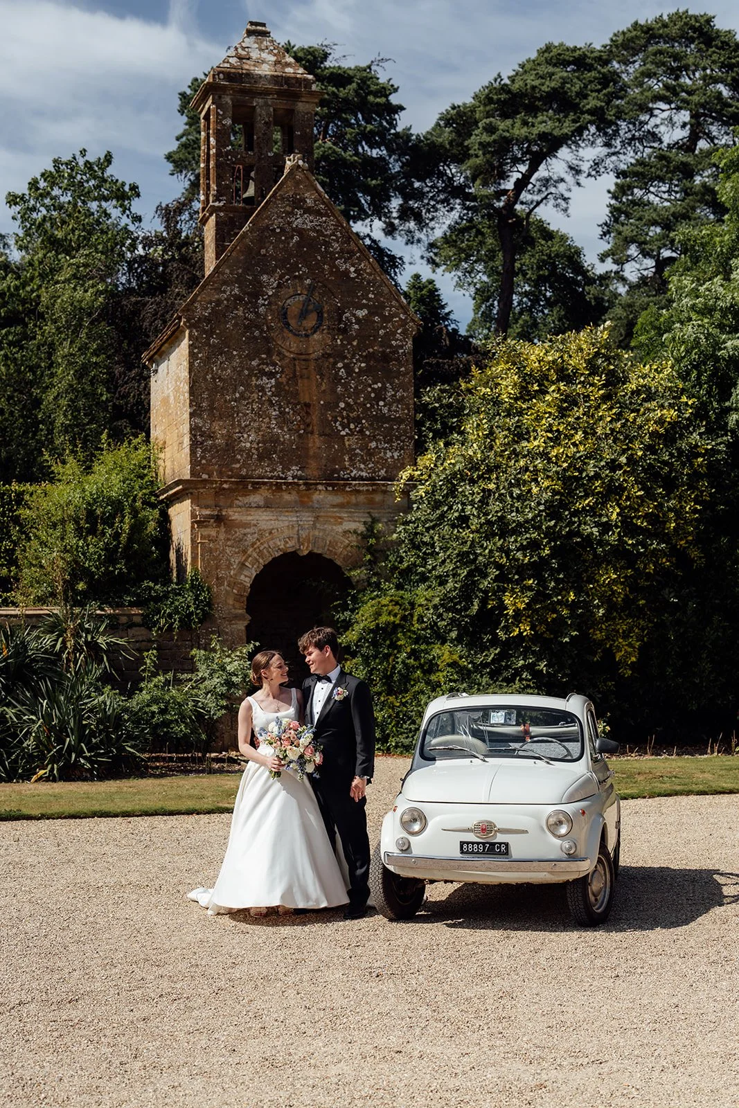 Rachel & Tom stand beside a small vintage white car outside the historic stone bell tower at Brympton House in Somerset, photographed by Just James Weddings.