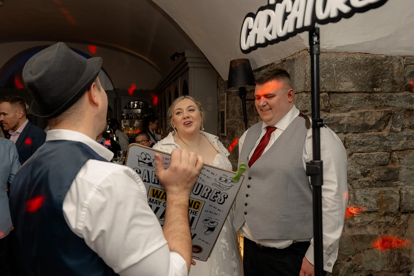 Becky & Rob laugh while watching a live caricature artist during their evening reception at Clearwell Castle in Gloucestershire, UK. Fun wedding entertainment moment with guests during their lively Clearwell Castle wedding party.