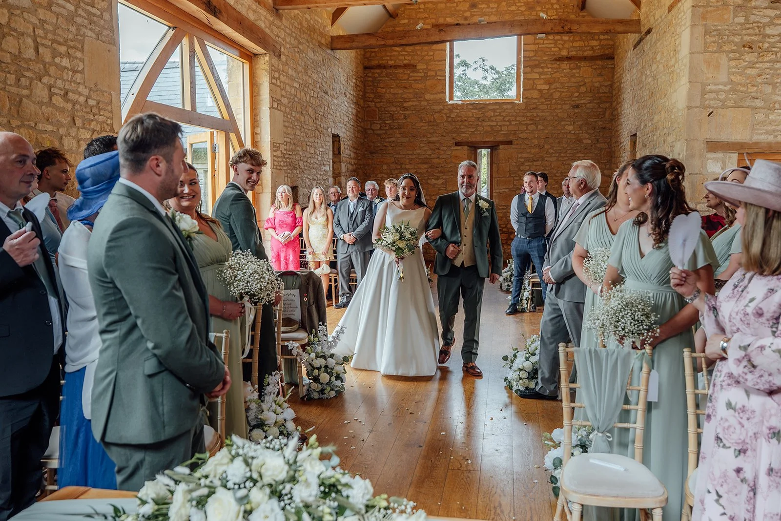 Chloe walking down the aisle with her father at The Barn at Upcote in the Cotswolds, surrounded by guests, photographed by Just James Weddings for natural ceremony moments.