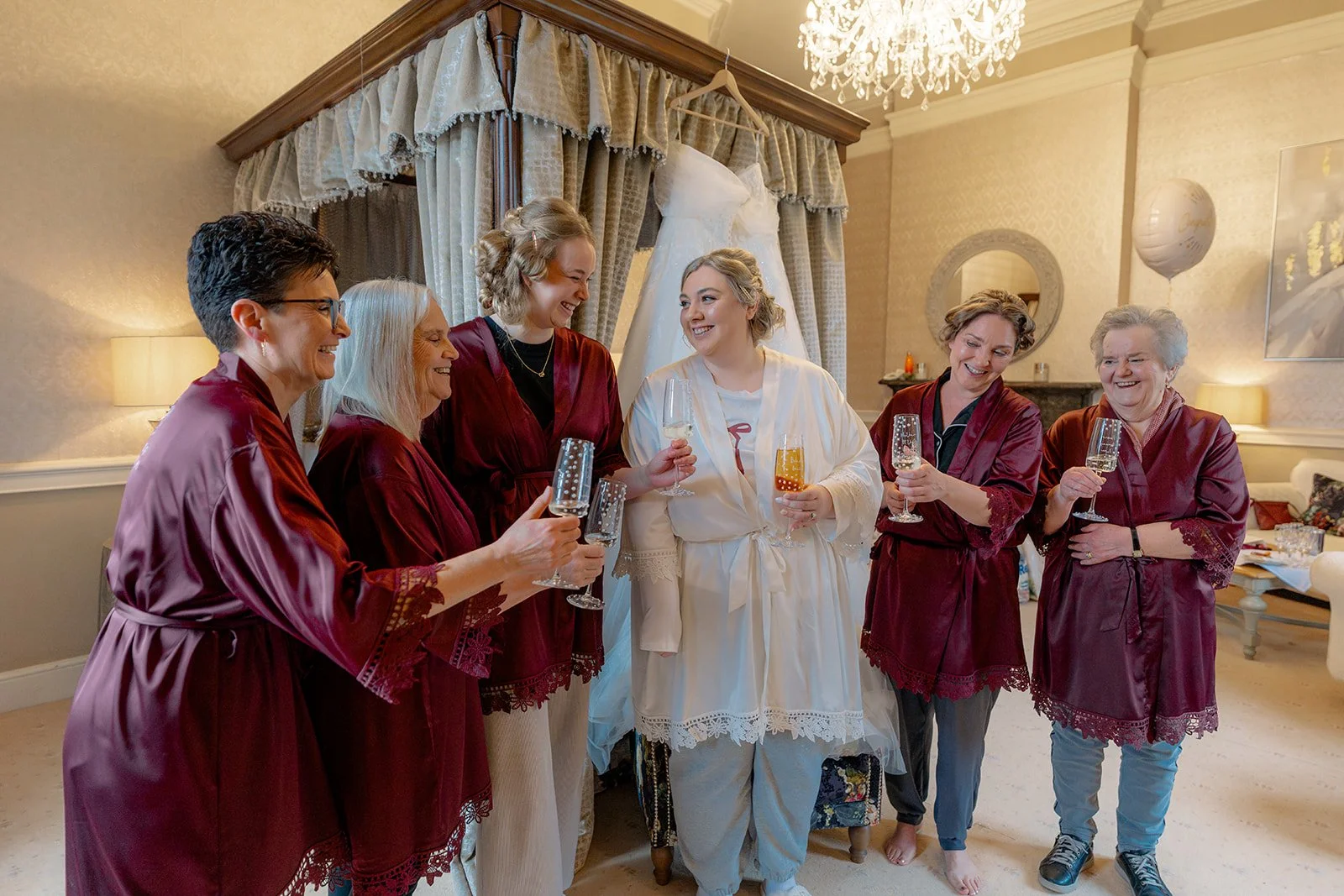 Becky & Rob celebrate with bridesmaids in burgundy robes, raising glasses and laughing in the bridal suite at Clearwell Castle, Gloucestershire, UK. Champagne toast before ceremony, joyful wedding morning prep and candid wedding photography.