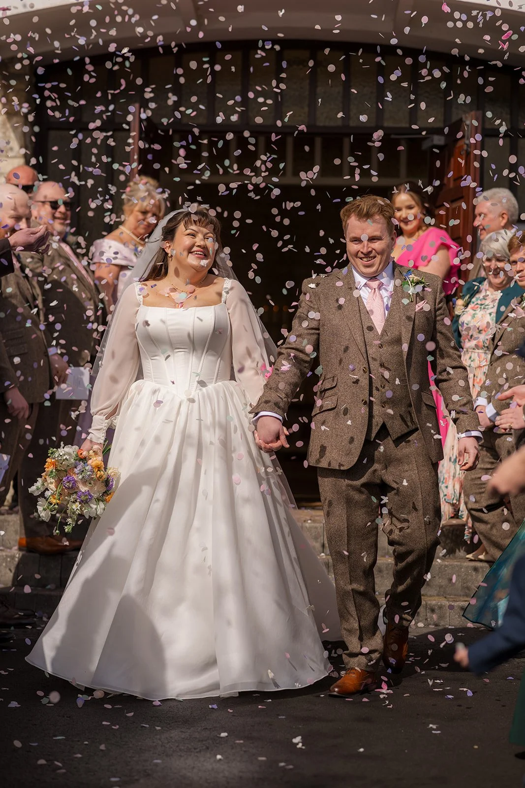 Emily & Josh walk through a joyful confetti throw outside Coombe Lodge Blagdon, Somerset, UK, holding hands and smiling as guests celebrate around them. This wedding photo captures the colour, energy and excitement of their Somerset wedding day.