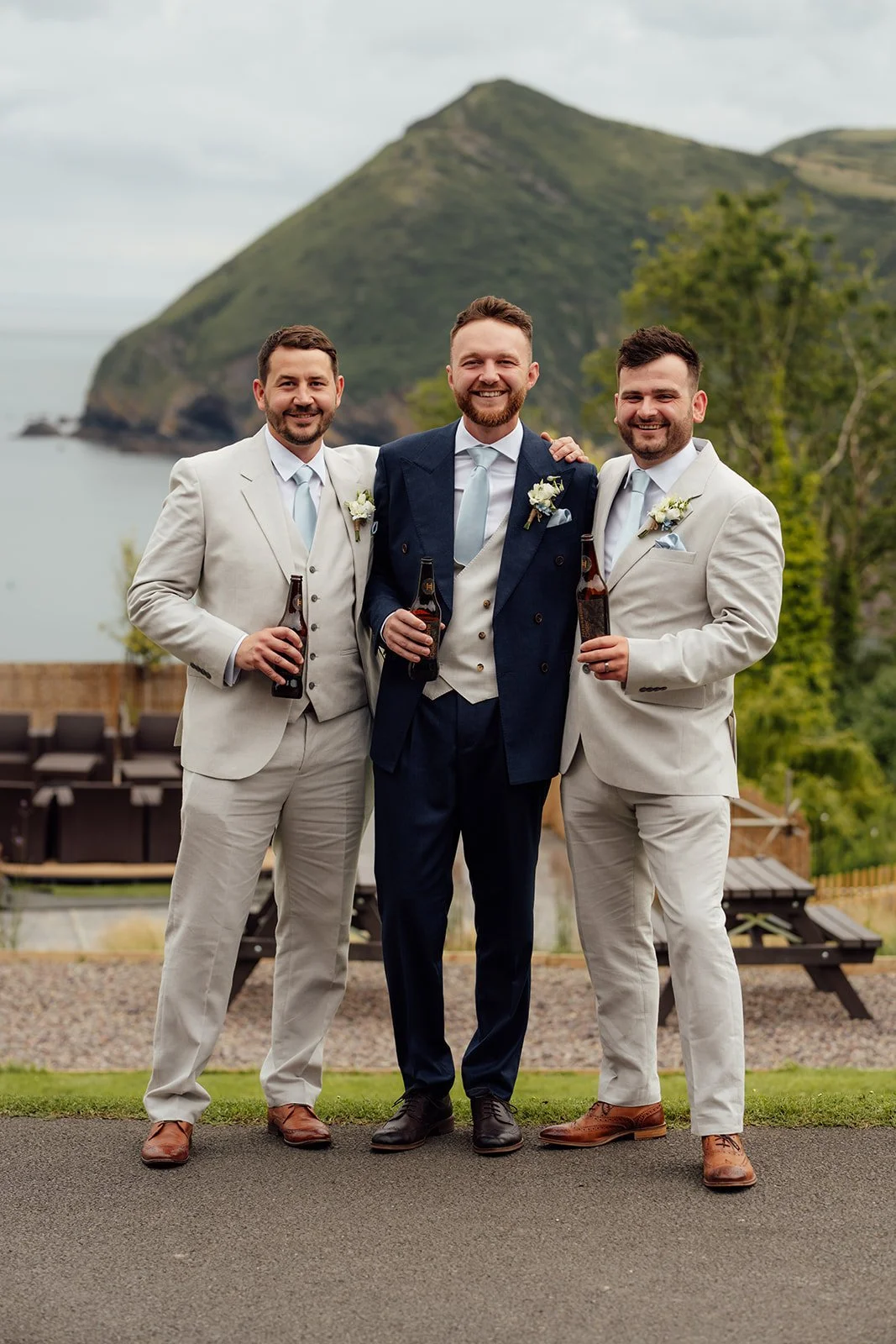 Connor stands smiling with his two groomsmen in coordinated navy and light suits, holding drinks with the dramatic North Devon coastline behind them at Sandy Cove Hotel, UK.
