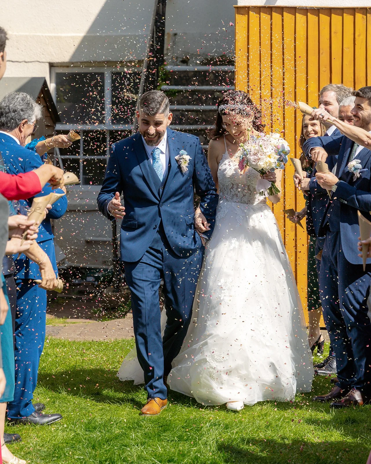 Ruth and Carmelo walk through a confetti shower after their wedding ceremony at No. 4 Clifton Village in Bristol, UK, surrounded by cheering friends and family.