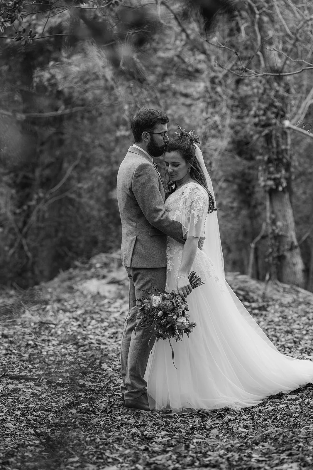 Black and white portrait of Maisie & Andrew embracing in autumn woodland at Stockbridge Farm Barn in Dorset, photographed by Just James Weddings for natural romantic wedding photography.