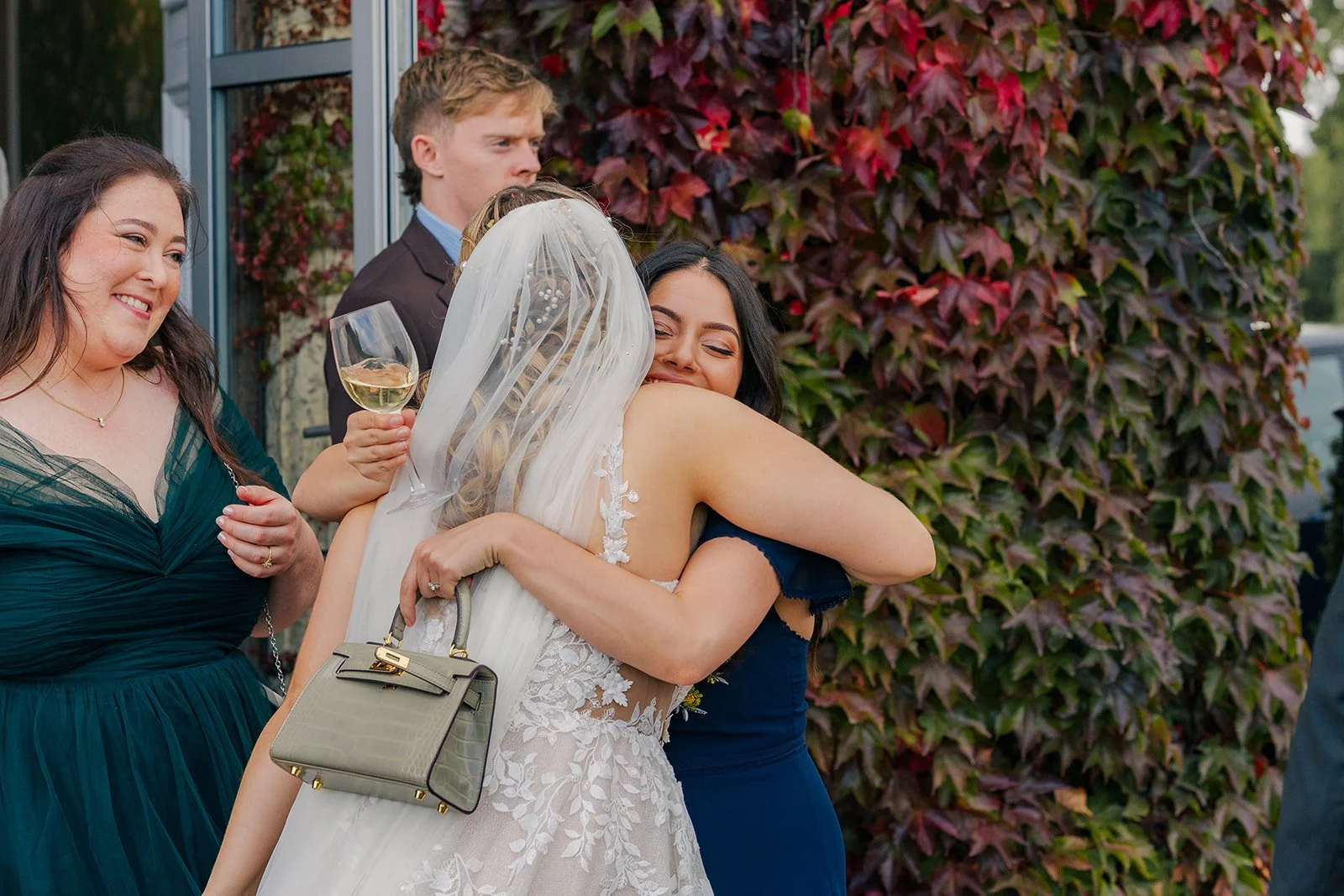 Lucy hugging a wedding guest outside Eastwood Park in Gloucestershire, with red ivy and guests nearby, photographed by Just James Weddings for candid wedding reception moments.