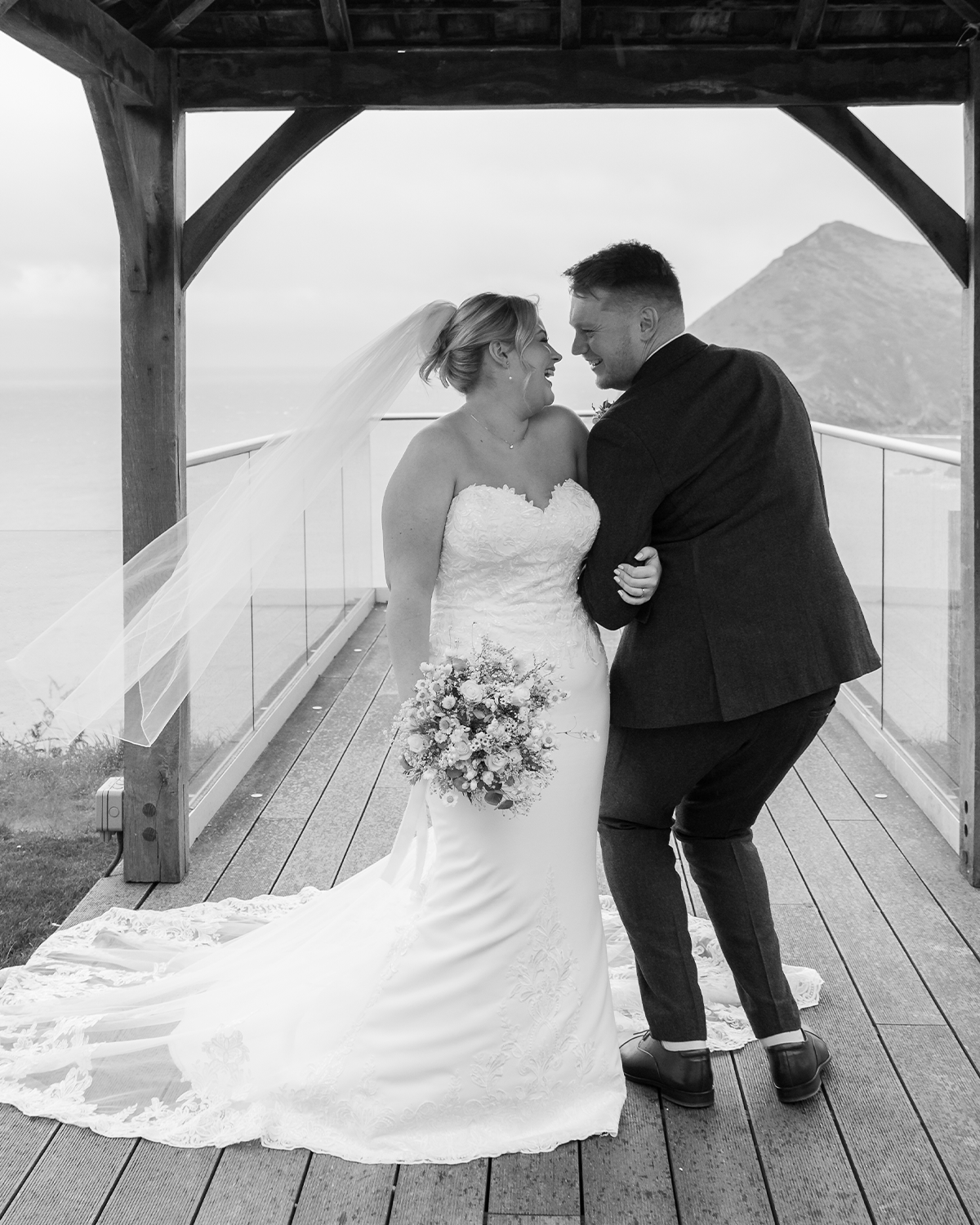 Black and white photo of a bride and groom smiling and holding each other on a wooden balcony overlooking the ocean with a mountain in the background, during their wedding.