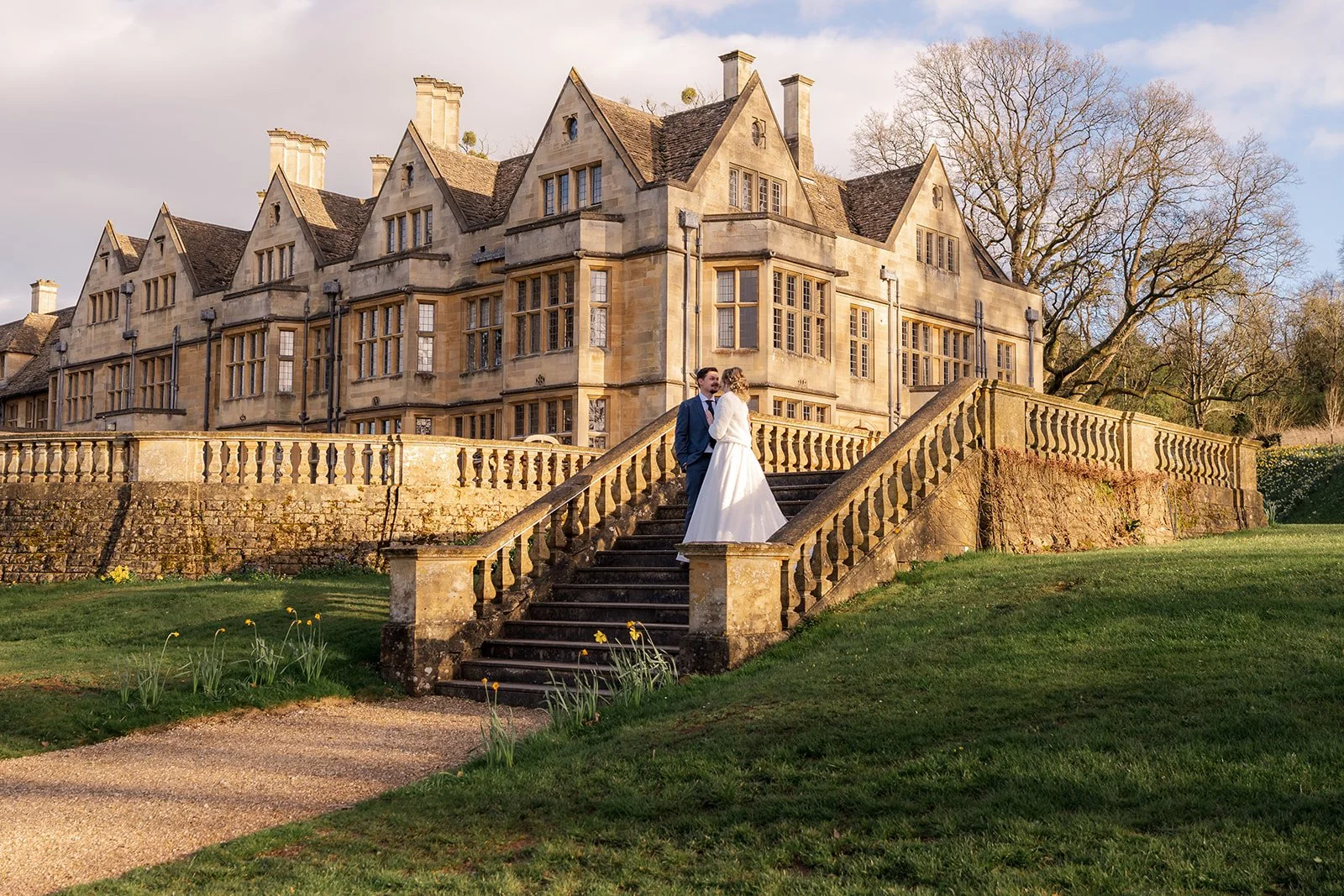 Jessie and Thomas stand together on the grand stone staircase at Coombe Lodge in Somerset, UK, with the historic country house creating an elegant backdrop for their wedding portrait.