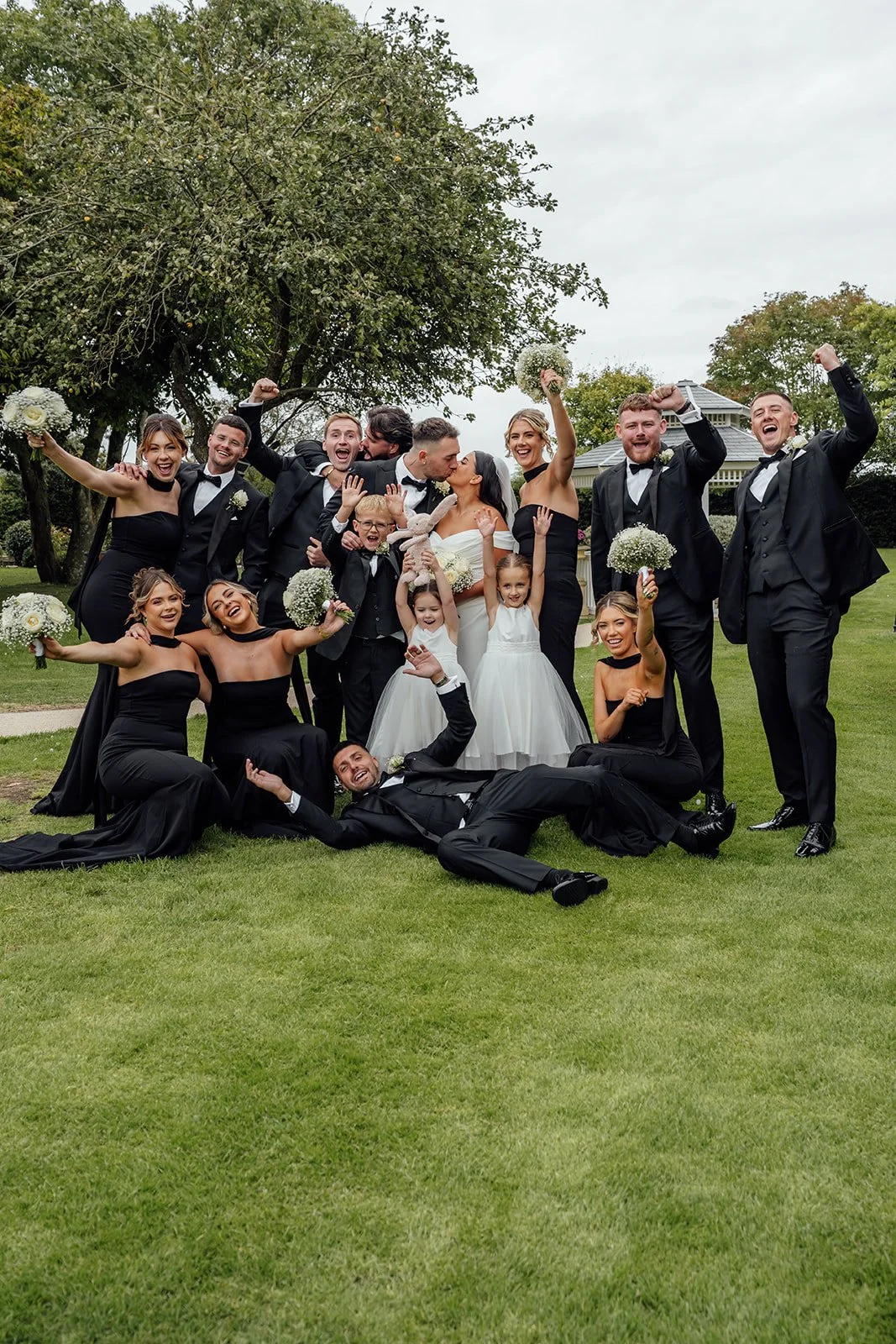 Nisha and Jack kiss in the middle of their wedding party at Batch Country House in Somerset, UK, surrounded by bridesmaids in black dresses, groomsmen in tuxedos, flower girls and page boys celebrating around them, by Just James Weddings.