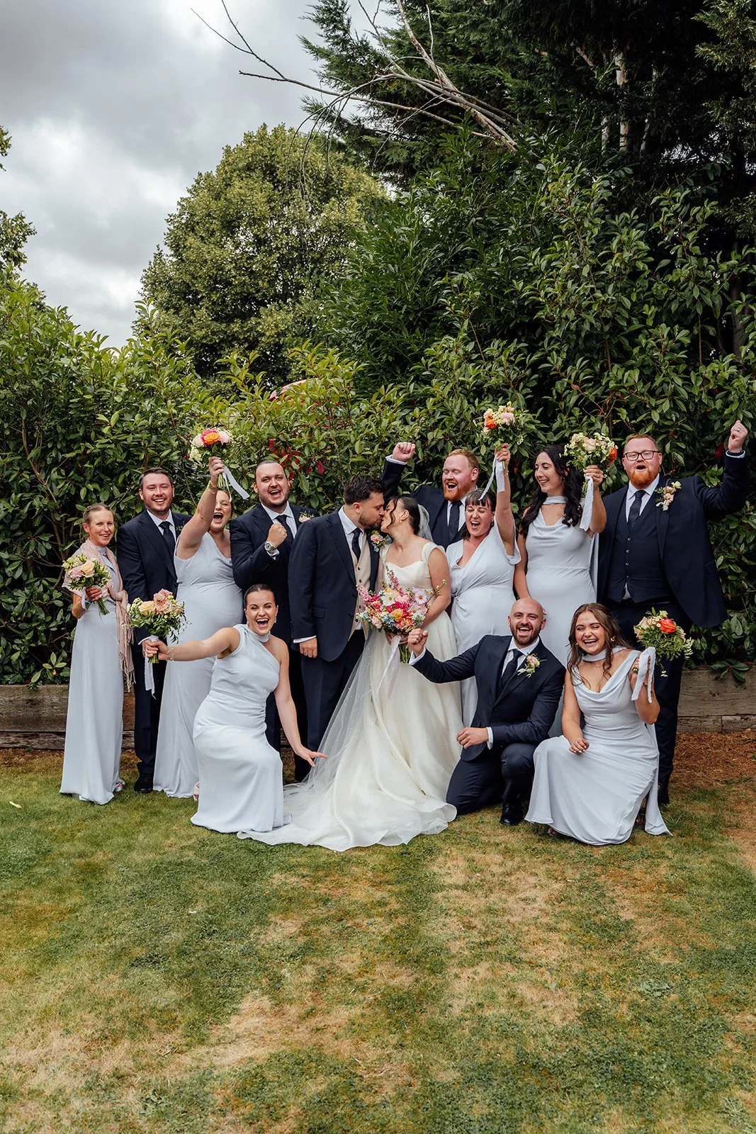 Harriet & Simon kiss in the centre of their cheering wedding party during a lively group photo at Dovecote Events in Oxfordshire, photographed by Just James Weddings.