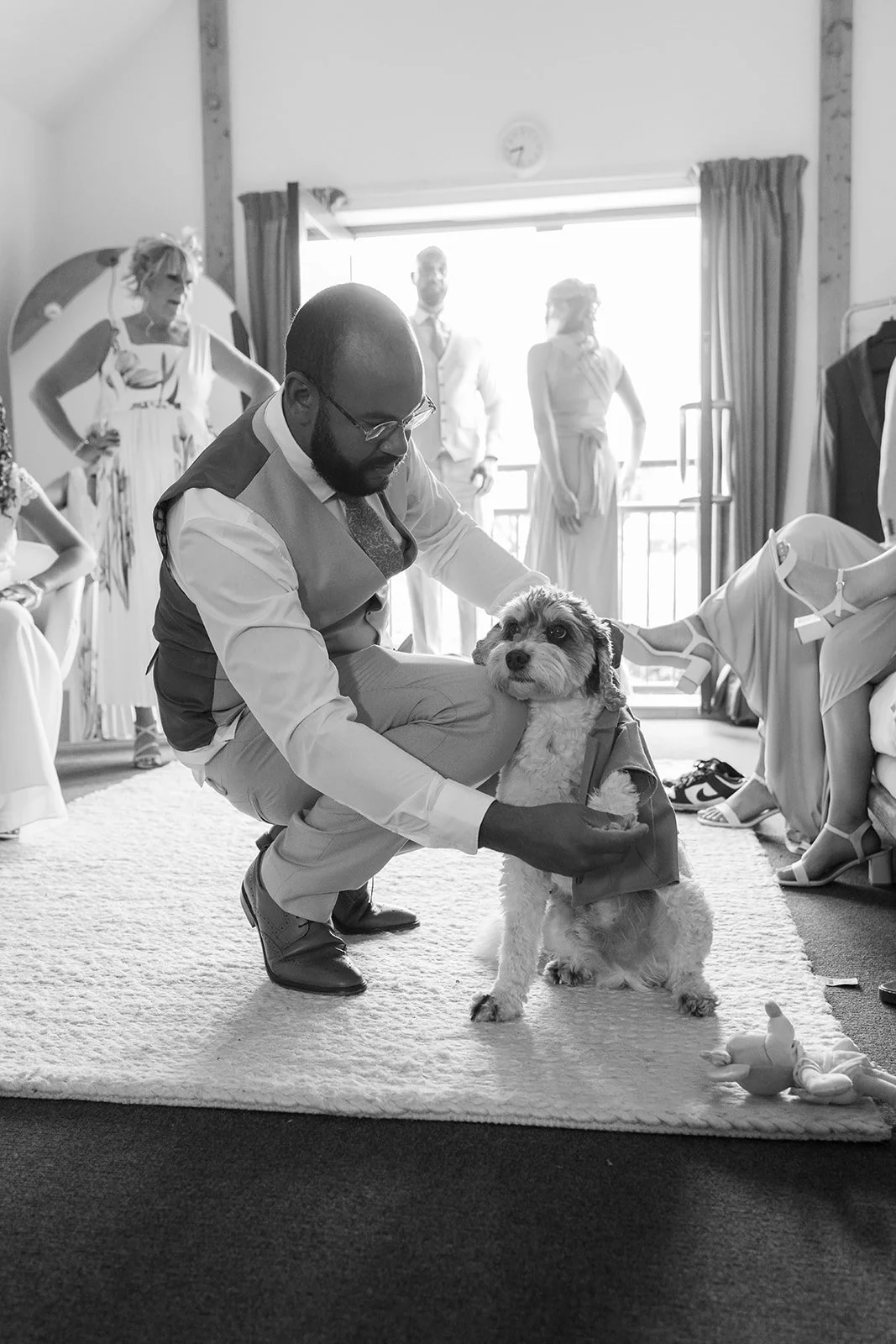 Black-and-white candid of Aaron crouching beside their dog Milo in his wedding outfit while guests gather during preparations at Rock Village Hall in Worcestershire, photographed by Just James Weddings.