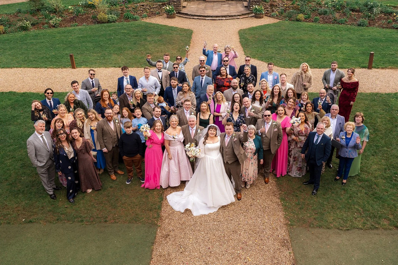 Emily and Josh stand at the centre of a large group wedding photo with friends and family in the gardens of Coombe Lodge in Somerset, UK.