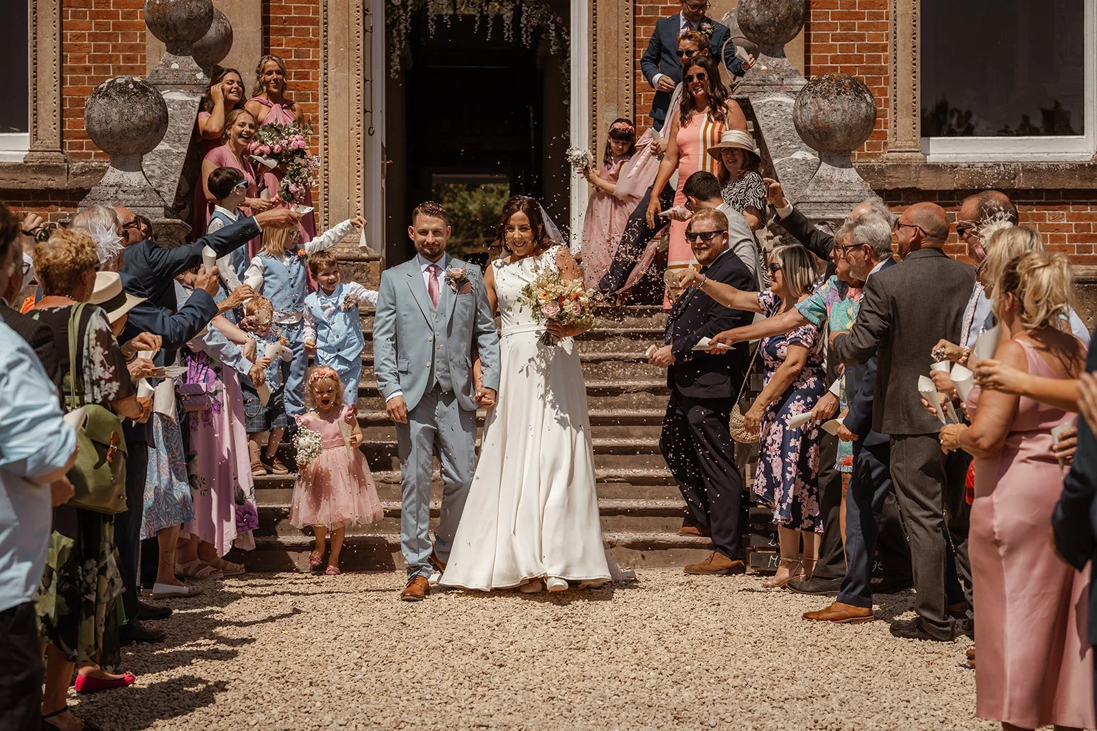 Michelle and Andy walk through a shower of confetti outside Crowcombe Court in Somerset, UK, surrounded by cheering friends and family after their wedding ceremony.