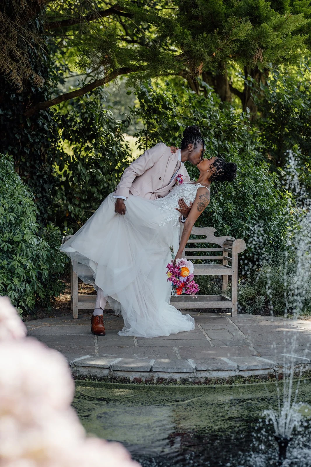 Kasey dipping Larisha for a kiss beside the garden pond during their wedding portraits at Oaks Farm Weddings in Surrey.