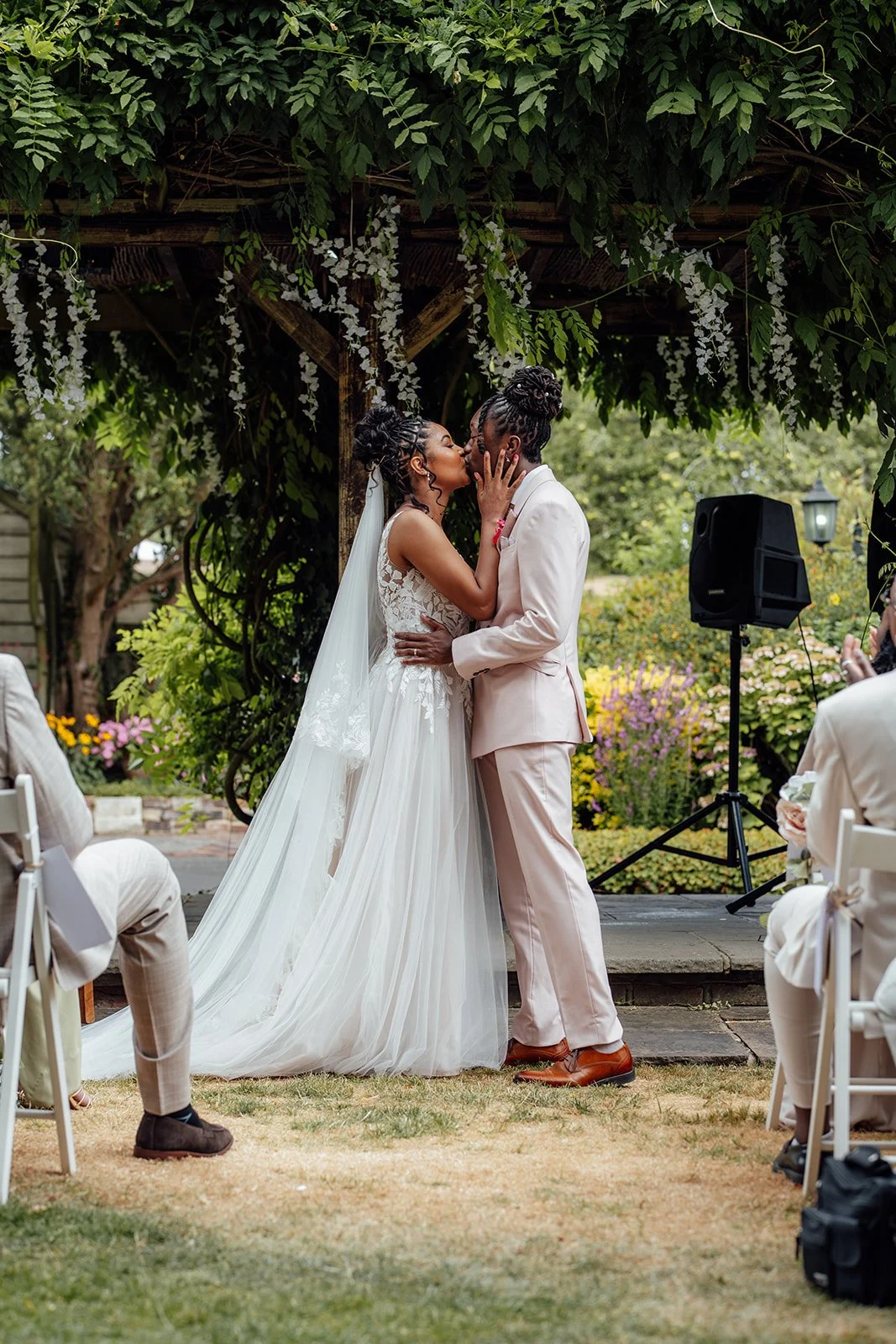 Kasey and Larisha sharing their first kiss beneath the leafy ceremony pergola during their outdoor wedding at Oaks Farm Weddings in Surrey.