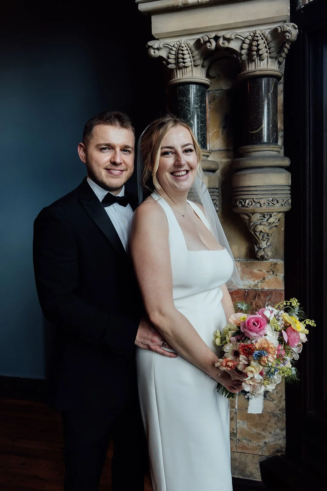 Bryony & Max smile together inside Insole Court in Cardiff, standing beside ornate stone columns and dark blue walls, with Bryony holding her colourful bouquet for a relaxed indoor wedding portrait that shows the venue’s historic charm well.
