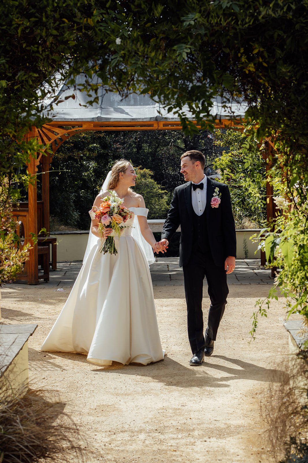 Caitlin and Kevin walking hand in hand through the gardens at Clevedon Hall in Somerset, smiling at each other beneath the leafy archway after their wedding ceremony.