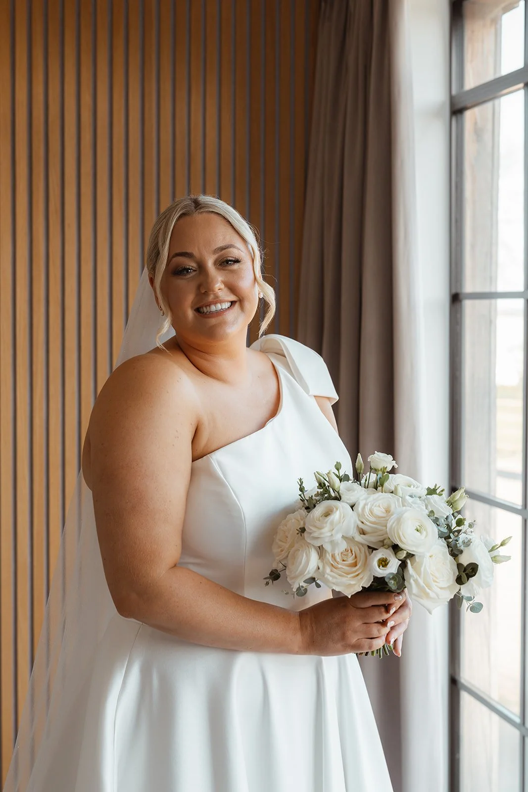 Jessie holding her bouquet and smiling beside a window during her wedding at Upton Barn & Walled Garden in Devon, UK. Soft natural light highlights her modern bridal gown, veil, and white florals ahead of Jessie & Jay’s ceremony.