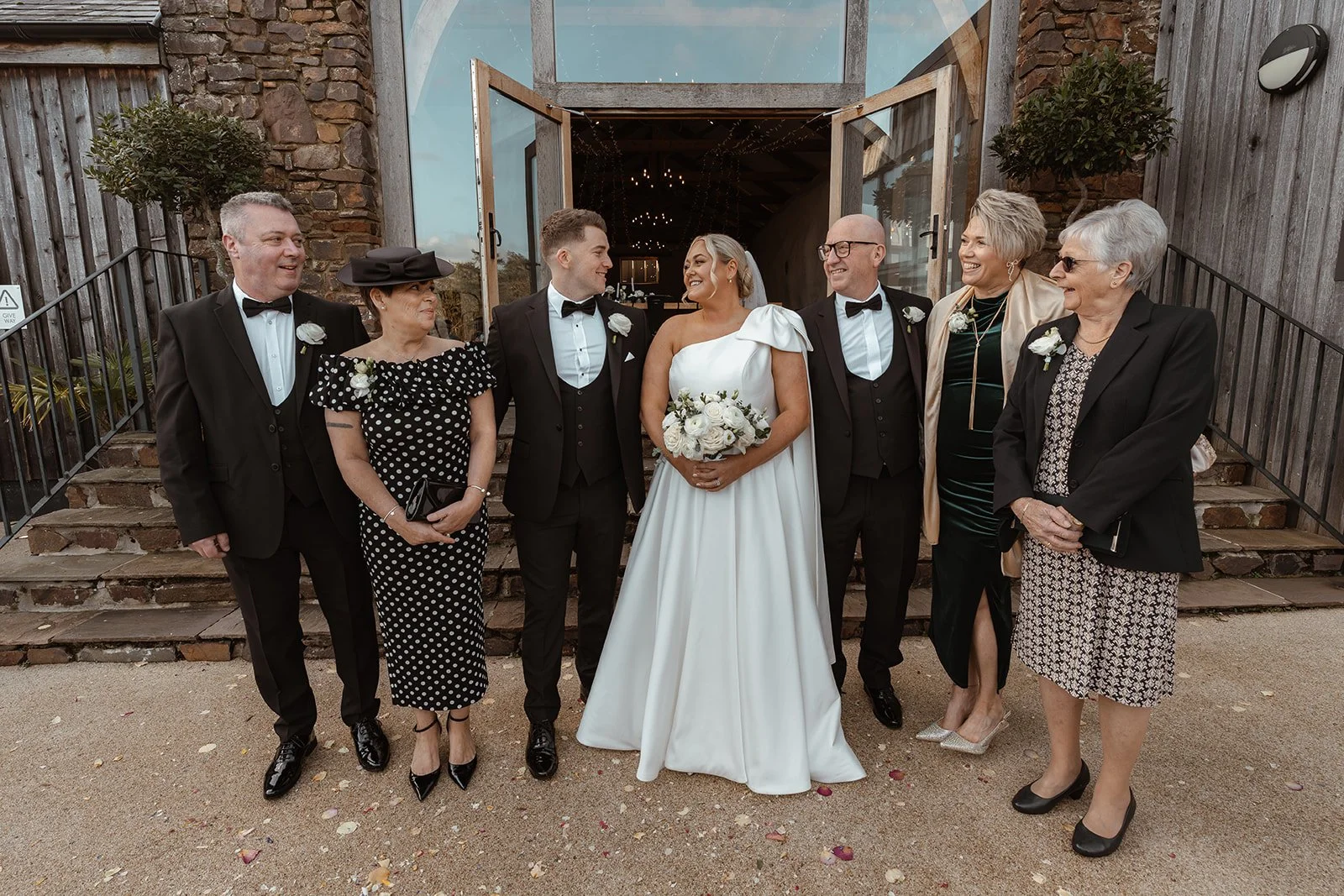 Jessie & Jay pose with family members outside Upton Barn & Walled Garden in Devon, UK. The group stands in front of the open barn doors, sharing laughter and connection in a warm, relaxed moment following the wedding ceremony.