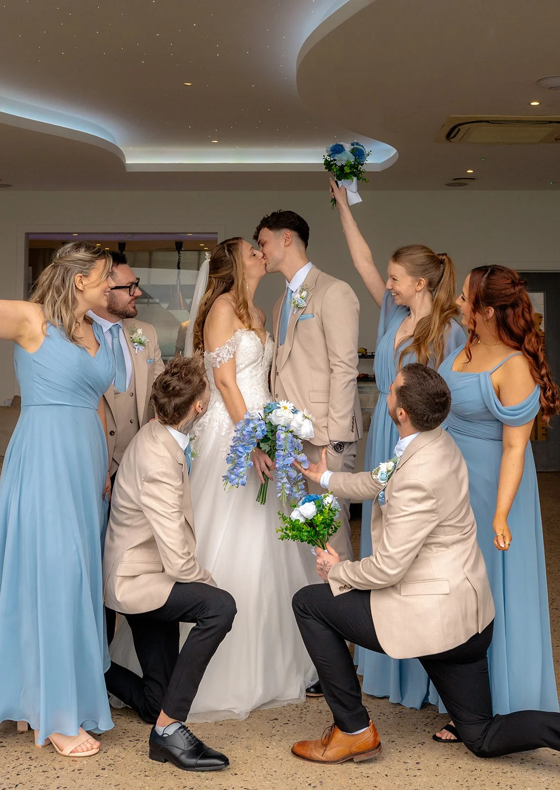 Hannah & Cameron share a kiss surrounded by their bridesmaids and groomsmen during fun wedding party portraits at Sandy Cove Hotel, North Devon. The wedding party celebrate around the couple holding blue bouquets in the bright reception space.