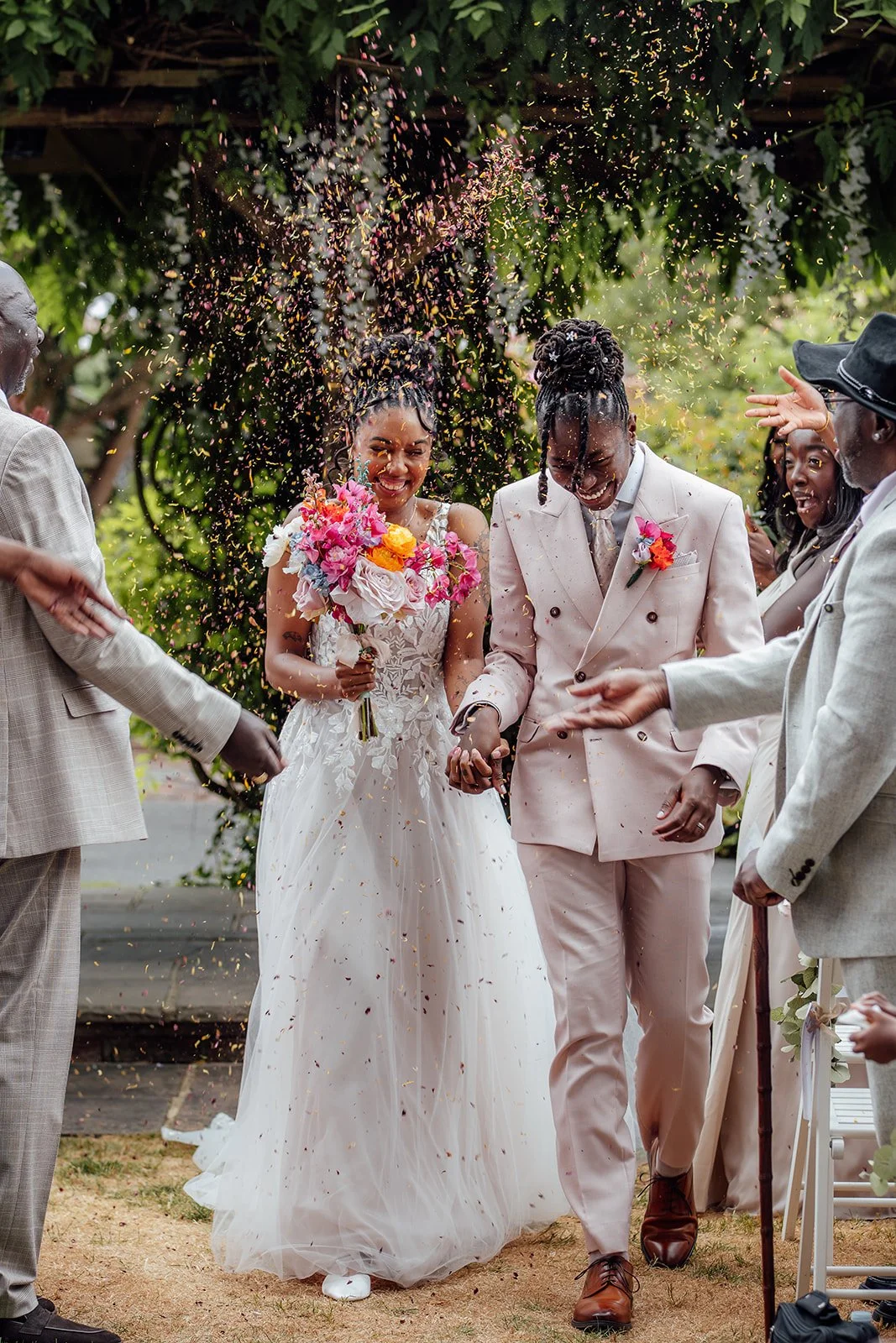 Kasey and Larisha walking hand in hand through colourful confetti after their ceremony at Oaks Farm Weddings in Surrey, smiling as guests celebrate around them.