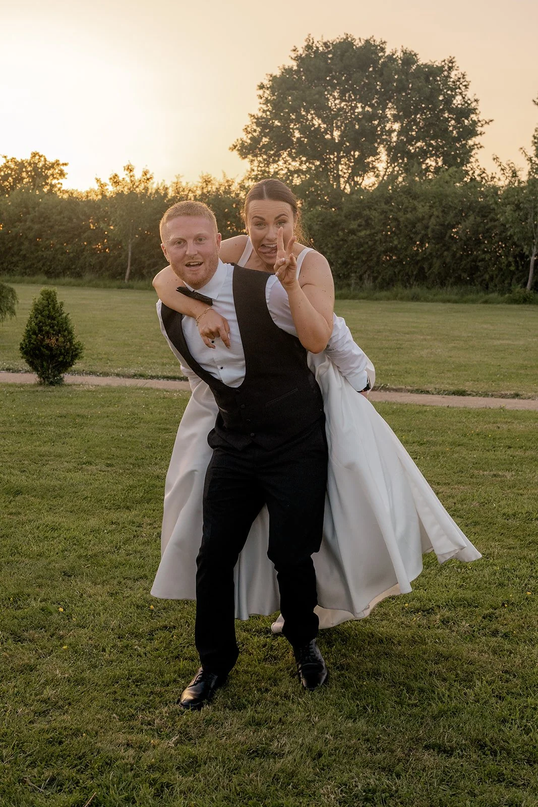 Playful golden hour photo of Lydia & Kieran on the lawn at Hencote Estate in Shropshire, UK, with Lydia riding on Kieran's back and making a peace sign during a relaxed, fun couple portrait after their wedding reception outdoors in summer.