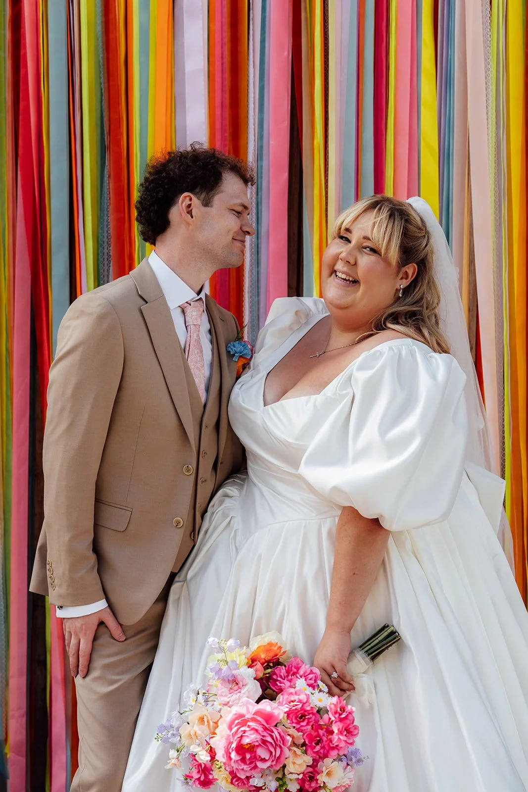 Holly & Alex pose in front of a colourful ribbon backdrop with Holly holding her bright pink bouquet at Sandy Cove Hotel in North Devon, photographed by Just James Weddings.