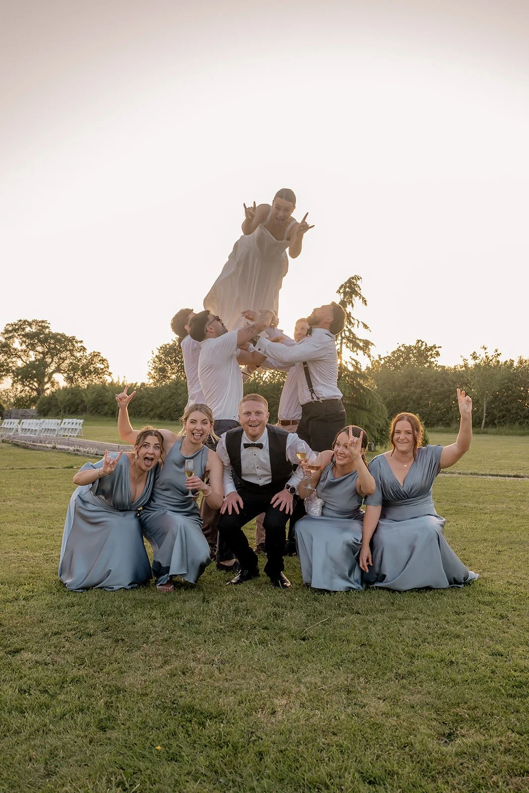 Fun wedding party photo of Lydia & Kieran with their bridesmaids and groomsmen on the lawn at Hencote Estate in Shropshire, UK, as one bridesmaid is lifted high in the air during a lively golden hour group portrait outdoors in summer.