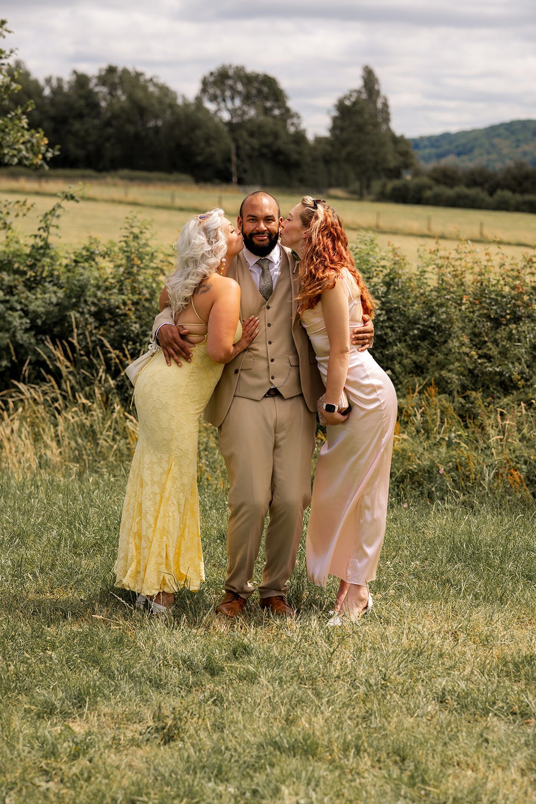 Aaron smiles as two wedding guests kiss his cheeks during relaxed countryside portraits at Rock Village Hall in Worcestershire, photographed by Just James Weddings.