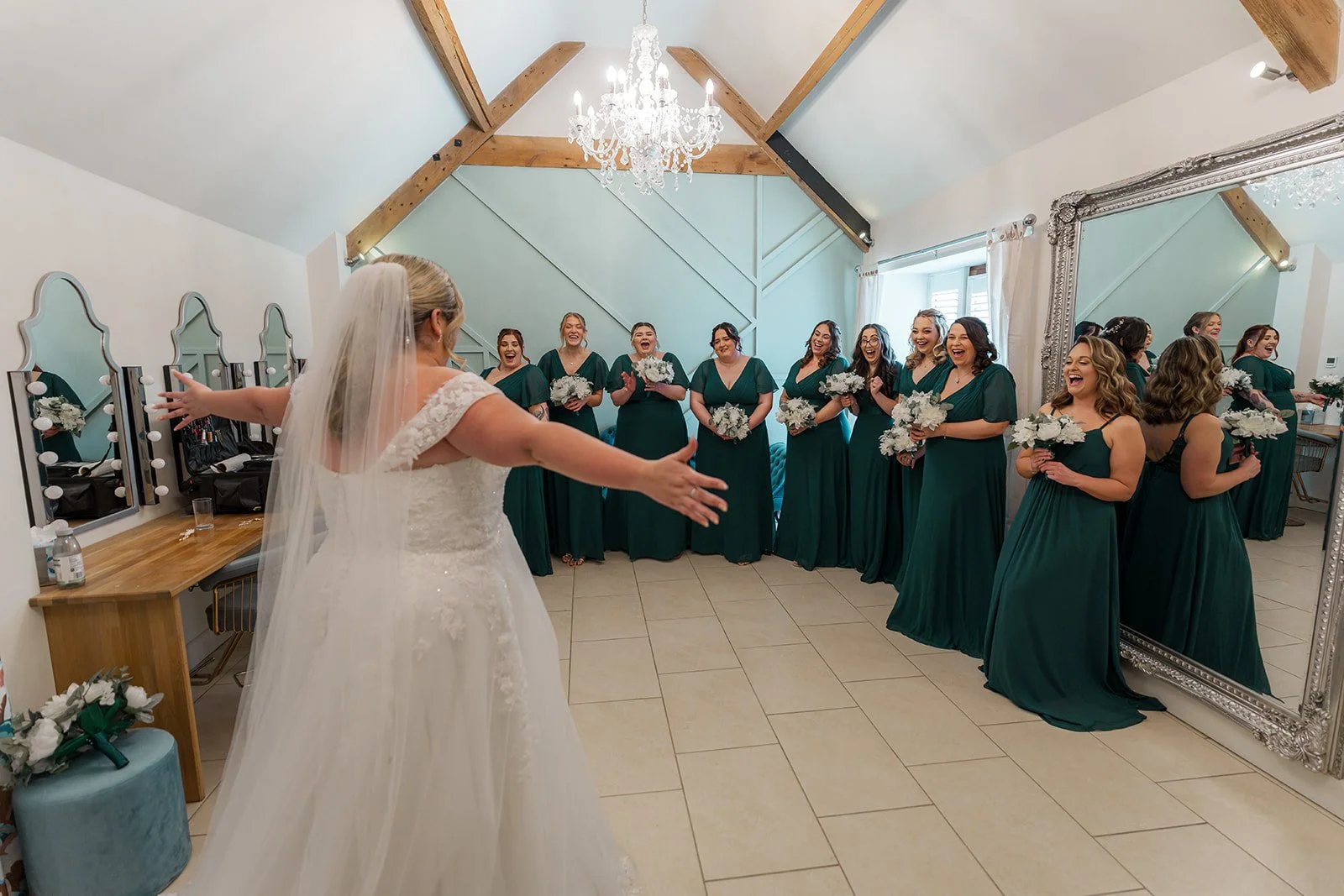 Bride revealing her wedding dress to bridesmaids in emerald green gowns inside Kingscote Barn, Cotswolds, UK, at Siobhan & Nicholas' wedding, a joyful bridal prep moment captured indoors in rustic barn wedding photography before the ceremony.