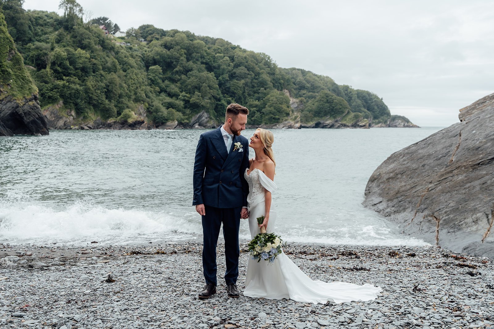 Amy & Connor stand together on a rocky beach beside the sea, surrounded by the North Devon coastline near Sandy Cove Hotel, UK.