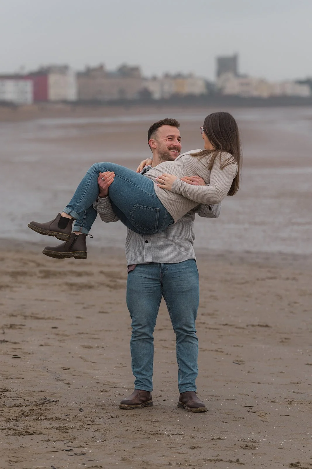 Emma & Dan share a joyful moment during their engagement and maternity shoot on the beach, with Dan holding Emma in his arms as they smile together. The soft shoreline and distant seafront buildings create a relaxed and romantic coastal portrait.