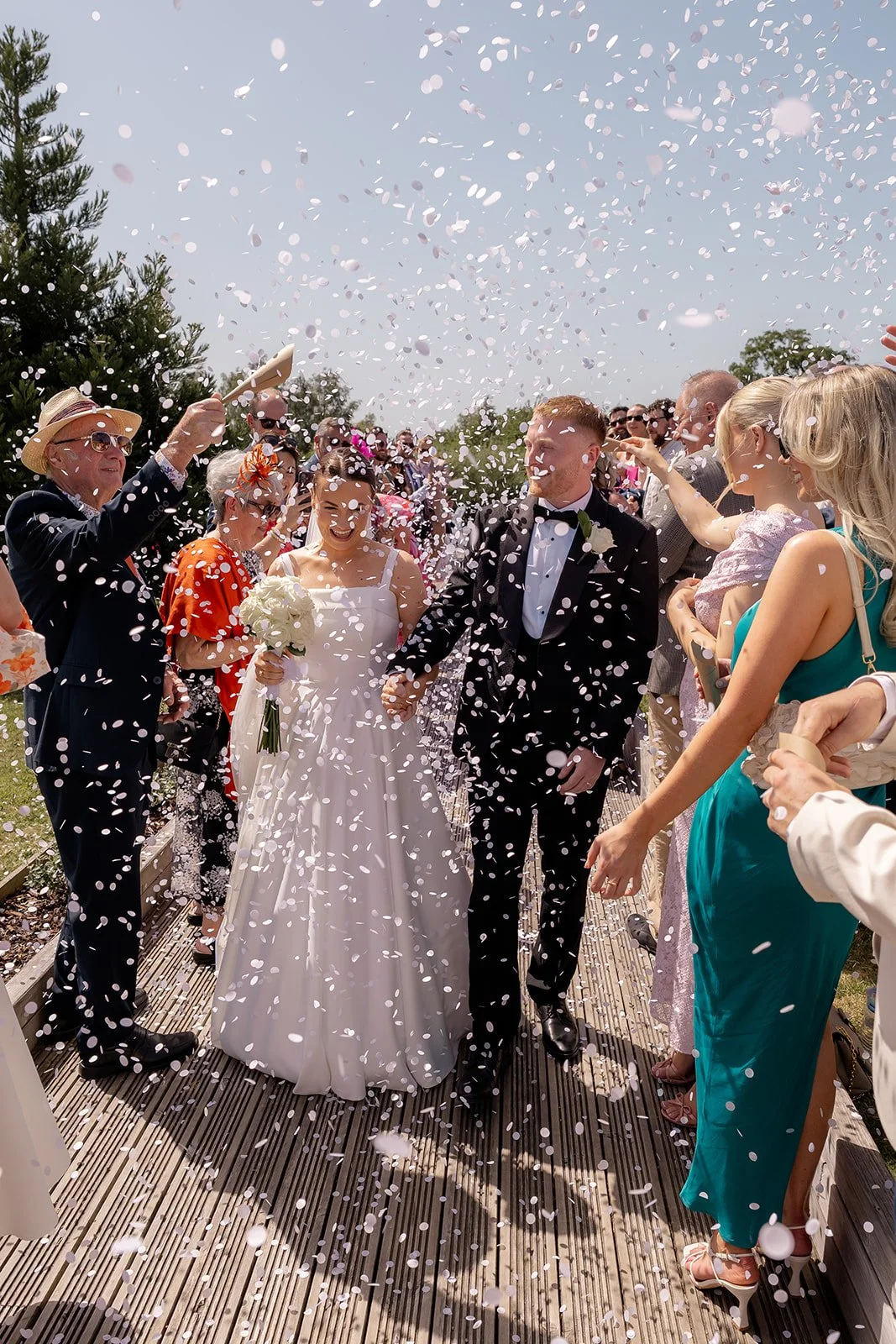 Confetti photo of Lydia & Kieran walking through cheering guests at Hencote Estate in Shropshire, UK, with white petals flying in bright summer light after their outdoor wedding ceremony and countryside celebration in the gardens.